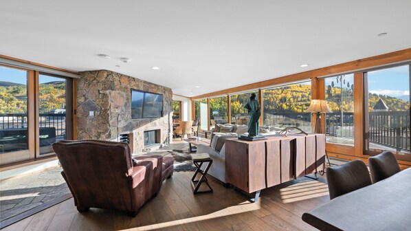 A spacious living area with a stone fireplace, leather furniture, and large windows offering a view of autumn foliage.