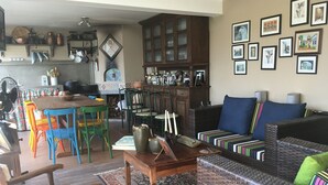 A cozy living room with a wicker sofa, colorful chairs, a dining table, and a kitchen area with copper pots. - Brotas Shangri-lá with 3 suites, pool and stunning views! (Caju)