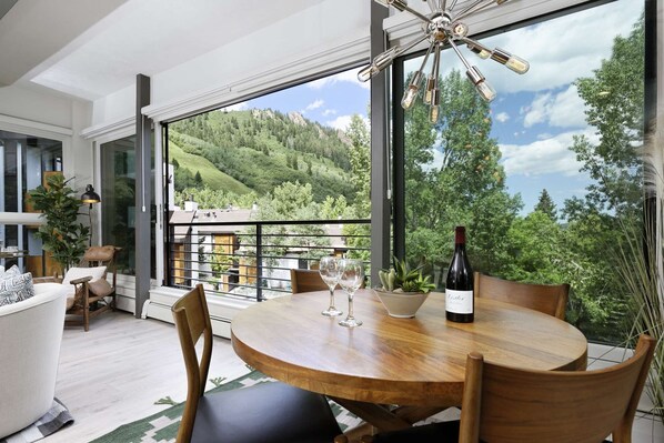 A dining area with a wooden table, chairs, and a bottle of wine. There is a view of a green landscape through large windows.