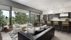 A modern living room with a black leather sofa, white armchairs, and a dining area with a round table. The kitchen features dark cabinets and a bar area with stools. Large windows provide a view of the outdoors. - Durant Condos by iTrip Aspen Snowmass (Aspen)