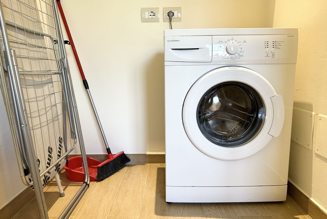 A white front-loading washing machine with a control panel, a metal drying rack, and a red-handled broom leaning against the wall.