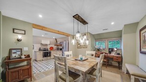 Un comedor con mesa y sillas de madera, una lámpara colgante y una cocina al fondo.