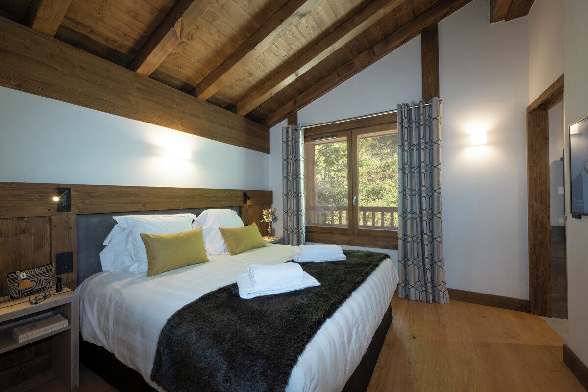 Un dormitorio con techo de madera, una cama grande con ropa blanca y un plaid oscuro, una mesita de noche con una lámpara, y una ventana con cortinas estampadas.