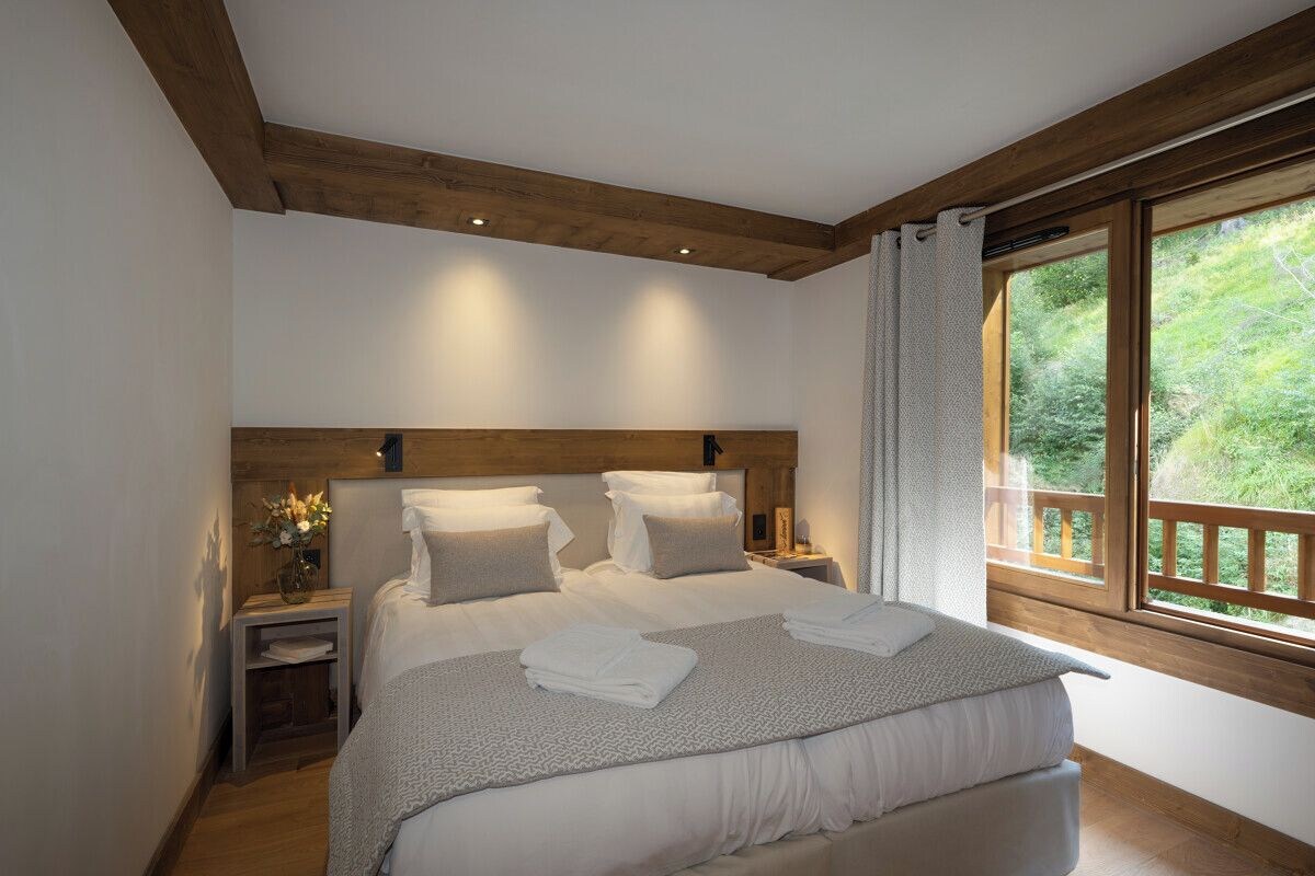 Un dormitorio con cabecera de madera, una cama grande con ropa de cama blanca, una mesita de noche con un jarrón de flores y una ventana con vista a follaje.