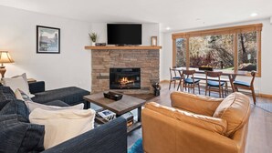 A living room with a stone fireplace, a flat-screen TV, a dining area with a table and chairs, and a large window with a view of trees.
