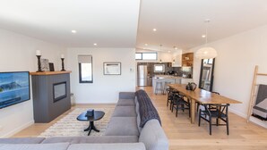 Una sala moderna con un sofá gris, una mesita de centro y un televisor sobre un mueble de madera. Se observa una cocina con comedor al fondo.