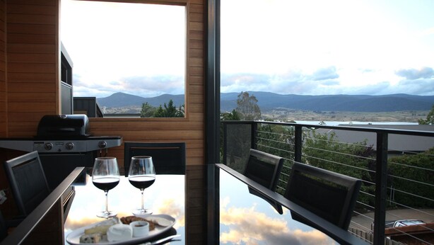 Un balcón con una mesa para dos, copas de vino, una parrilla y una vista panorámica de montañas y un lago.