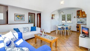 A living room with a sofa, a dining table, and a kitchen area with a microwave and a TV. - VILLA FARO (Cap d´Artruix)