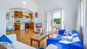 A modern living room with a sofa, coffee table, and a kitchen area with cabinets and a TV. - VILLA FARO (Cap d´Artruix)