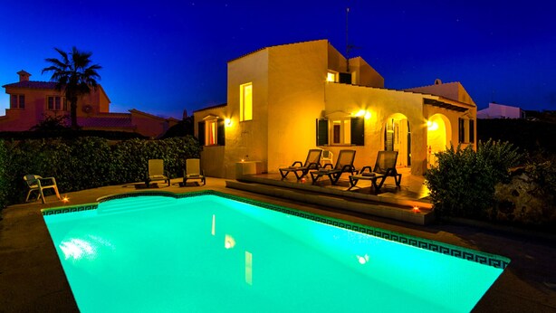 A swimming pool with lounge chairs and a building with warm lighting. - VILLA FARO (Cap d´Artruix)