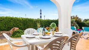 An outdoor dining area with white tables and chairs, a view of a lighthouse, and a well-maintained hedge in the background. - VILLA FARO (Cap d´Artruix)