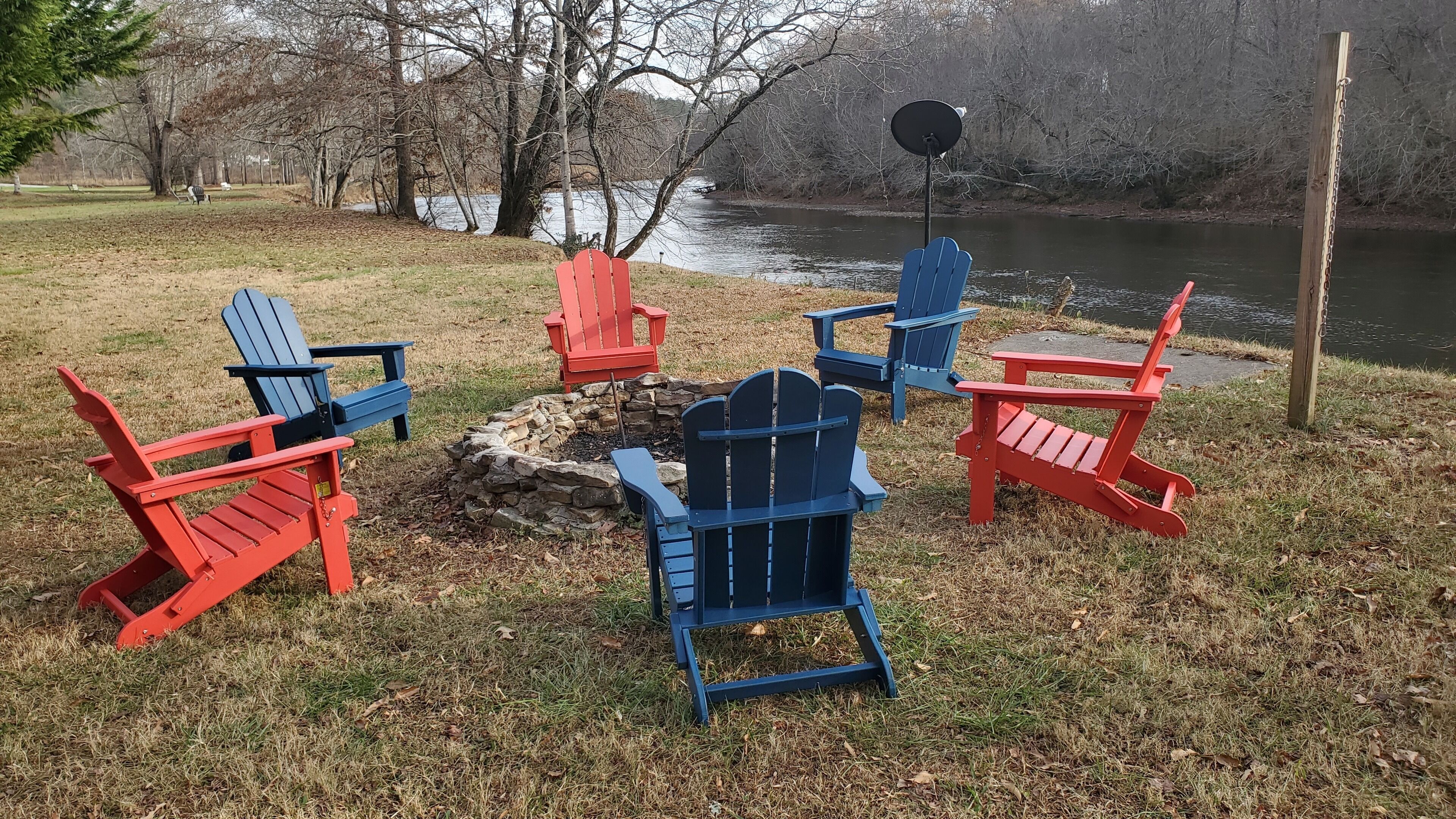 Un groupe de chaises Adirondack est disposé autour d’un foyer dans un parc, avec un lac en arrière-plan.