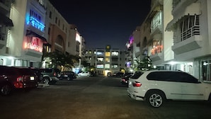 A nighttime view of a parking lot with several cars parked. The buildings on either side have illuminated signs and windows.