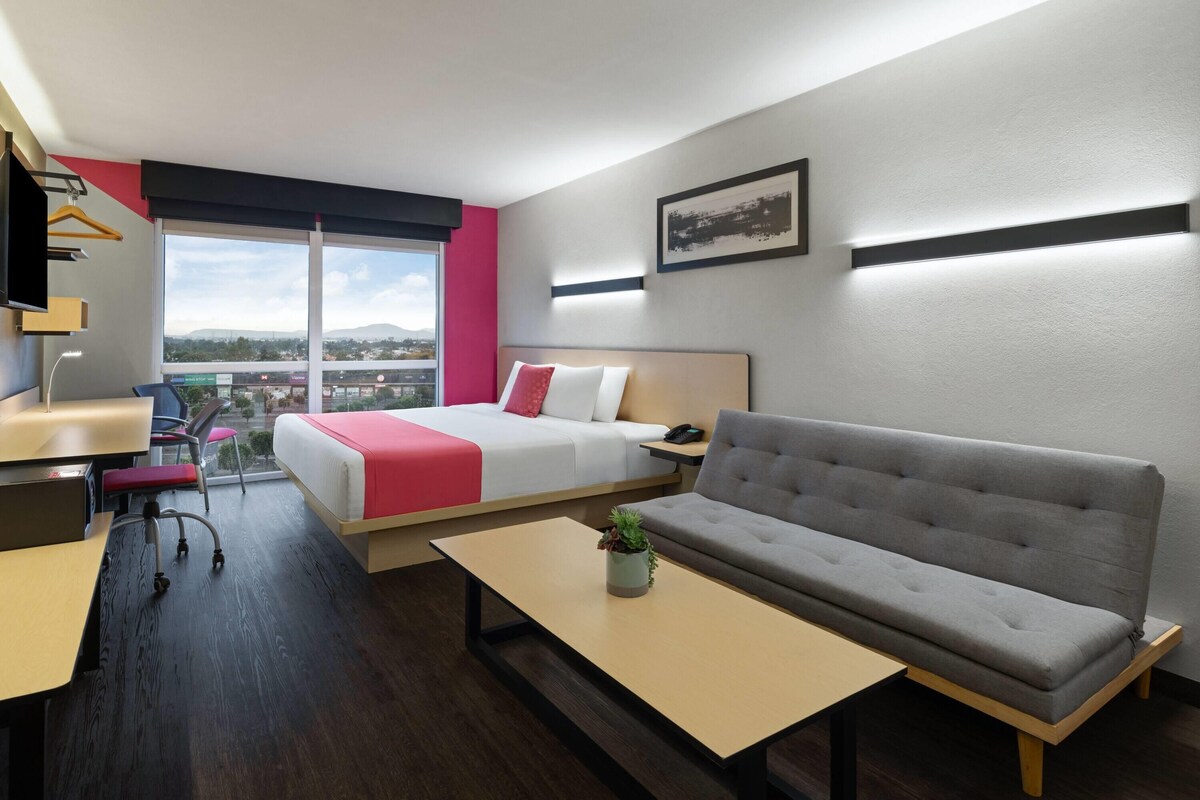 Ett modernt hotellrum med en säng, soffa, skrivbord och utsikt över staden.