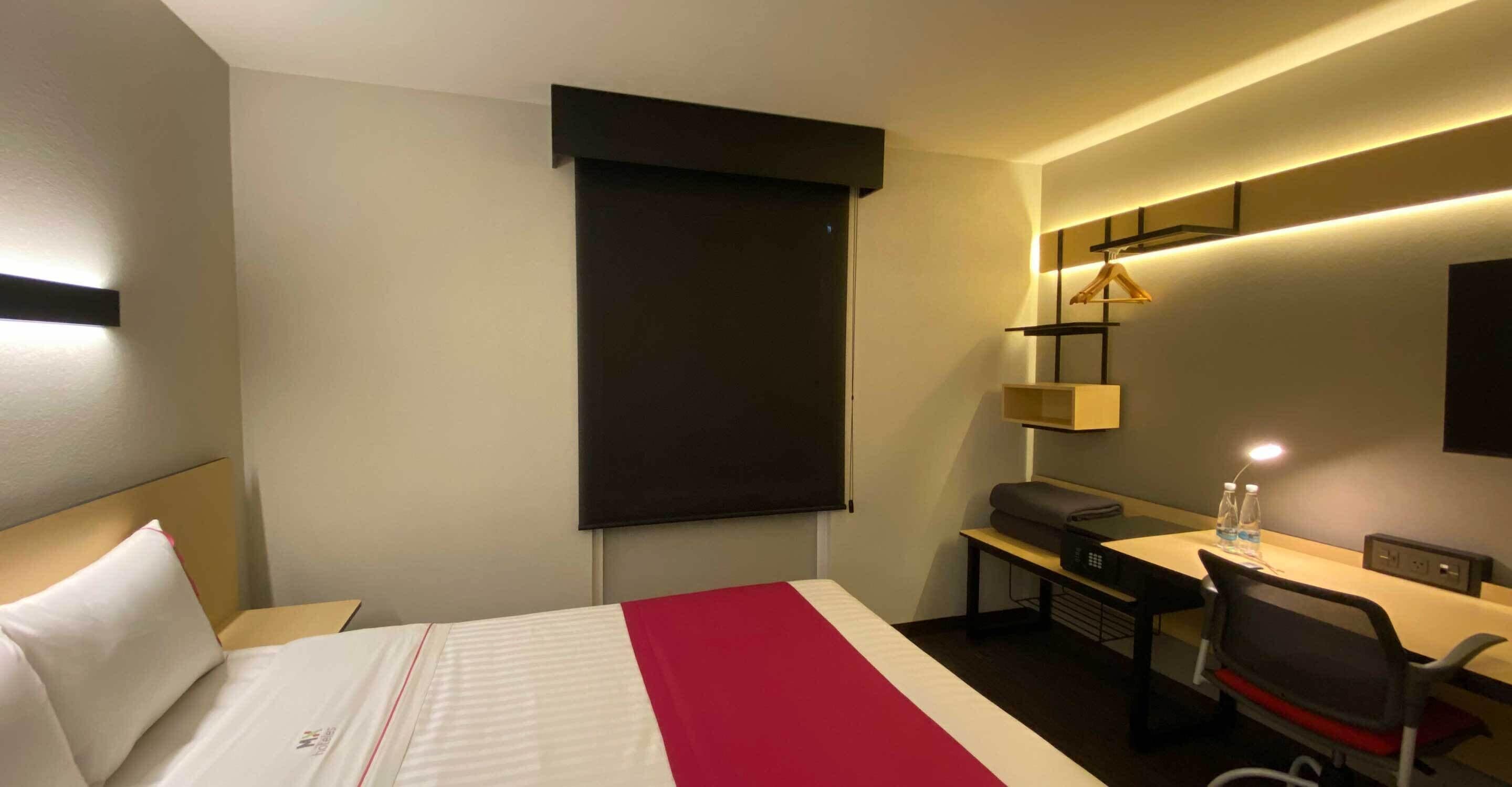 A hotel room with a bed, a desk, a chair, a TV, and a bedside table with a lamp and water bottles.