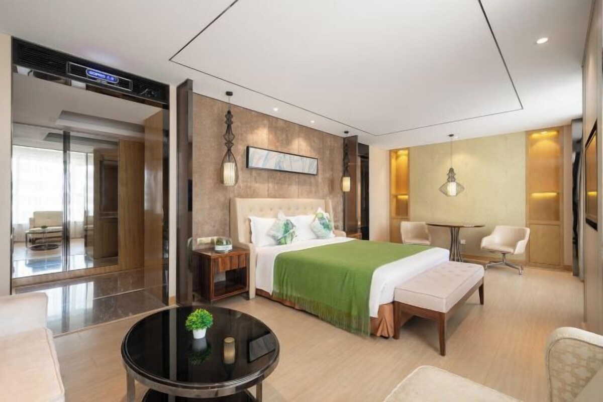 A modern hotel room with a large bed, a bedside table, a round coffee table, and a seating area with a chair and a small table.