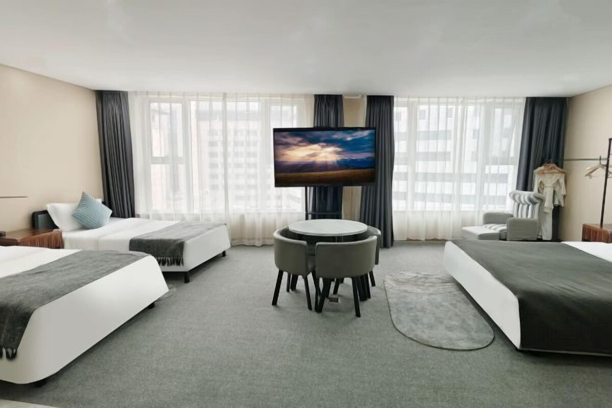 A hotel room with a bed, a desk with chairs, a television, and a view of the cityscape through large windows.