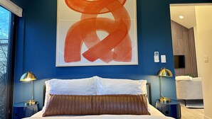 A bedroom with a large abstract painting, a bed with white and brown bedding, two bedside lamps, and a view of the outdoors through a window. - Greenwood Getaway - Modern and New, Walk to Shops (Seattle)