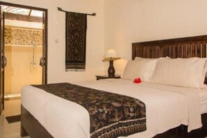 A hotel room with a bed, a nightstand with a lamp, a shower area, and a wall hanging. - Lily Lane Villas (Ubud)