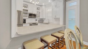 A kitchen with white cabinets, a stainless steel refrigerator, a microwave, and a sink. There are three wooden stools with cushioned seats. - Award-Winning Resort Amenities | Grand Pavilion 67 (Isle of Palms)