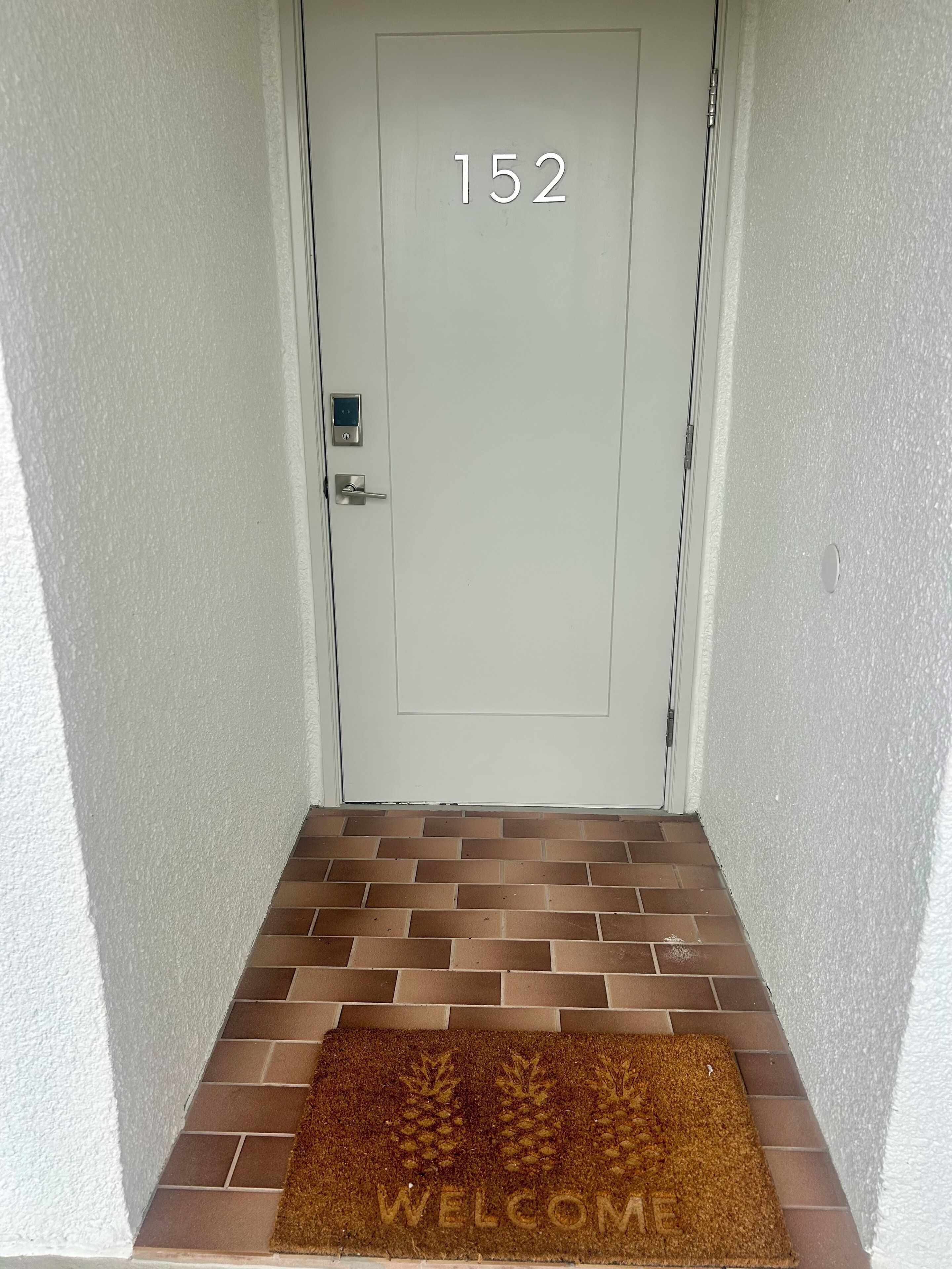 A white door with the number 152, a digital lock, and a brown doormat with 'WELCOME' engraved.