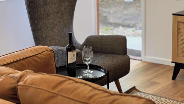A modern living room with a grey upholstered armchair, a brown leather sofa, a small black table with a wine bottle and glasses, and a view of the outdoors through a glass door. - 3 Bed Budget Brisbane Gem With Parking Near Gabba (Woolloongabba)