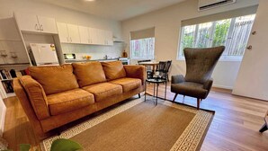 A modern living room with a sofa, armchair, and coffee table. There is a kitchen area with cabinets and a refrigerator. The room has large windows and a view of greenery outside. - 3 Bed Budget Brisbane Gem With Parking Near Gabba (Woolloongabba)