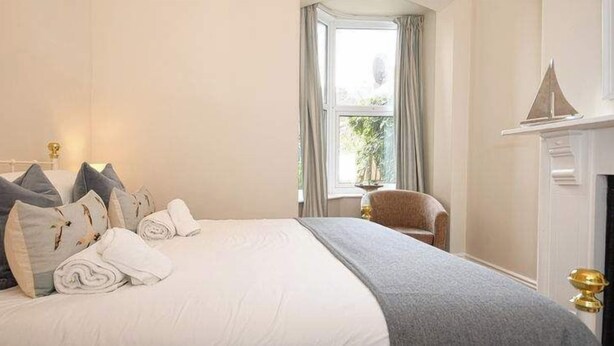 A neatly made bed with pillows and a blanket, a chair, a window with curtains, and a fireplace with a mantel. - Ground Floor At The Lawns - Lovely chic ground floor apartment (Filey)