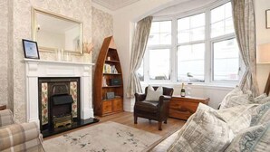 A living room with a fireplace, a television, a bookshelf, and a window with curtains. - Ground Floor At The Lawns - Lovely chic ground floor apartment (Filey)