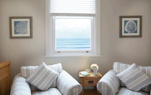A room with two striped sofas facing a window with a view of the ocean. There are two framed pictures on the walls, a small wooden nightstand with a book and a cup, and a wooden cabinet to the left.
