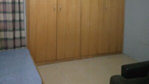 A bedroom with a large wooden wardrobe, a bed with a checkered bedspread, and a plain wall. - Short Stay Apartment (Accra)