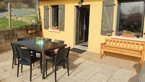 Une maison jaune avec une terrasse en bois, une table avec des chaises et un banc.