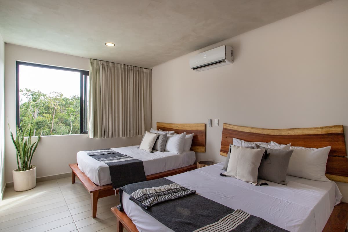 Habitación de hotel con dos camas, una planta en maceta, un ventanal grande y un equipo de aire acondicionado.