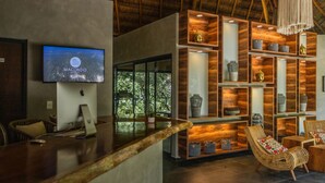 A modern interior with a wooden reception desk, a Macondo TV screen, and a wall of shelves with decorative items. - Macondo Tulum  (Tulum)