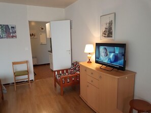 A room with a television on a wooden cabinet, a chair, a small table with a lamp, and a door leading to another room.