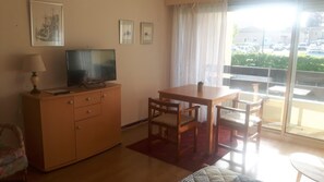 A living room with a wooden cabinet, a television, a dining table with chairs, and a large window with a view of parked cars.
