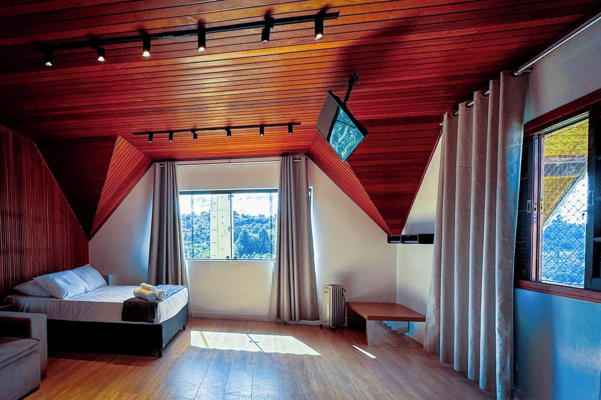 Een slaapkamer met een schuine kap, een bed, een bankje, een televisie en grote ramen.