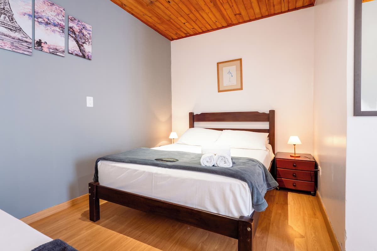 Een netjes opgemaakt bed met een houten hoofdbord, een nachtkastje met een lamp en een muur met ingelijste kunstwerken.