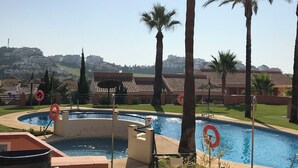 Una piscina con palmeras, un puesto de socorrista y vistas de edificios al fondo.