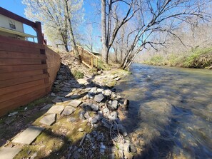Eine Holzterrasse neben einem klaren Bach, mit einem Steinpfad, der zu einer Holzbrücke führt.