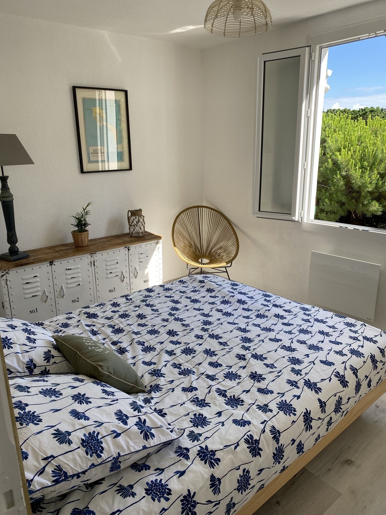 Quarto com cama, poltrona, cômoda, luminária e janela com vista para vegetação.