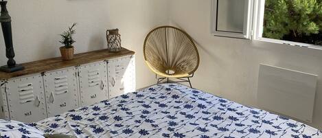 Quarto com cama, poltrona, cômoda, luminária e janela com vista para vegetação.