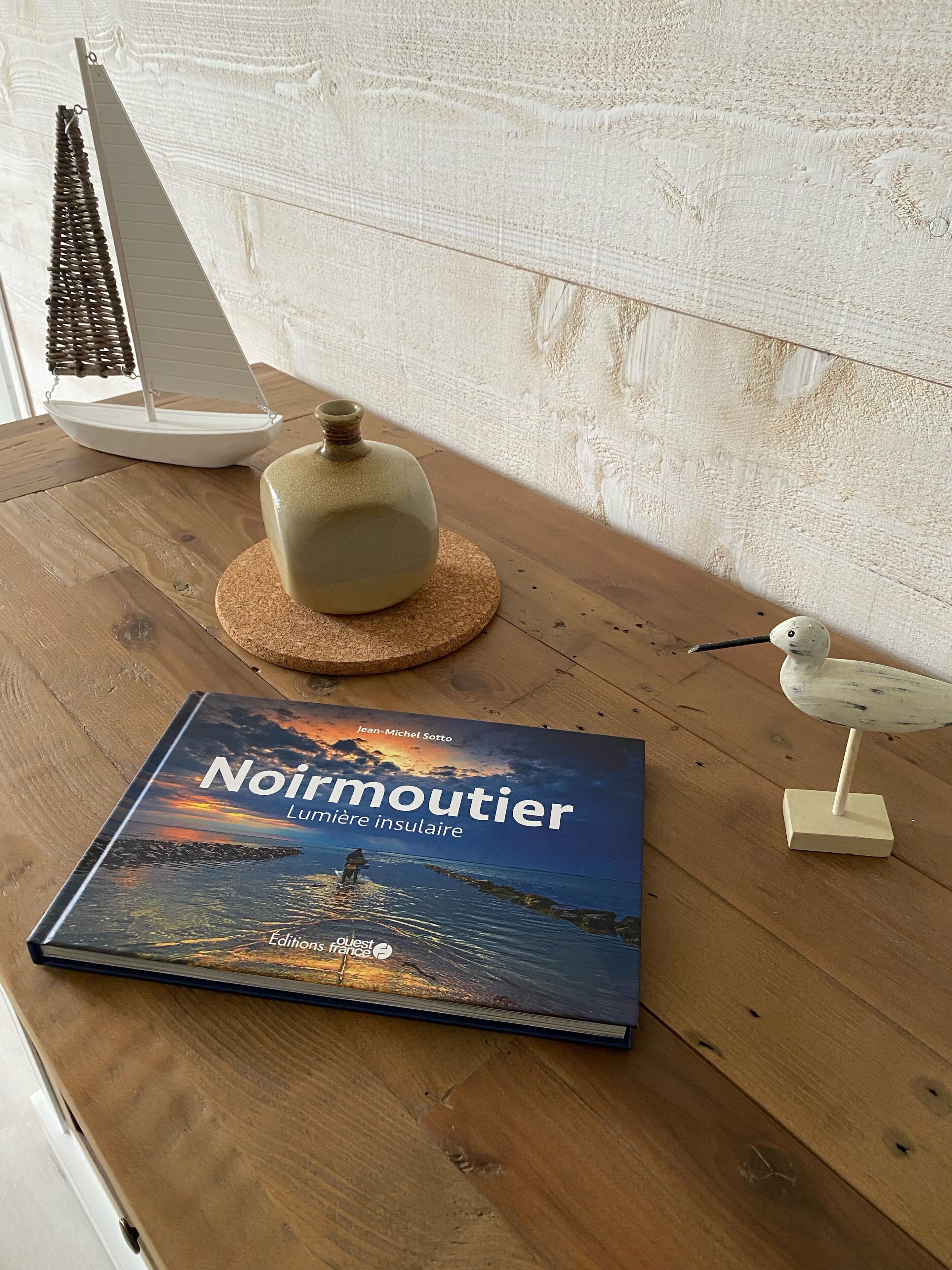 Un livre intitulé « Noirmoutier » posé sur une surface en bois, un vase en céramique, une petite maquette de voilier et une figurine d’oiseau.