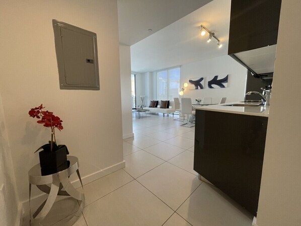 A modern kitchen with a dark countertop and white cabinets, a white dining area with a white sofa, and a living room with a white sofa and a large window.