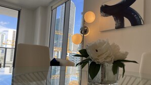 Une chambre moderne avec une table en verre, un vase rempli de fleurs blanches et une vue sur de hauts immeubles, visible à travers de grandes fenêtres.