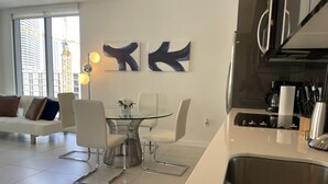 Une cuisine moderne dotée d’un évier en acier inoxydable, d’un coin repas avec une table en verre et des chaises blanches, et d’œuvres d’art abstraites accrochées au mur.