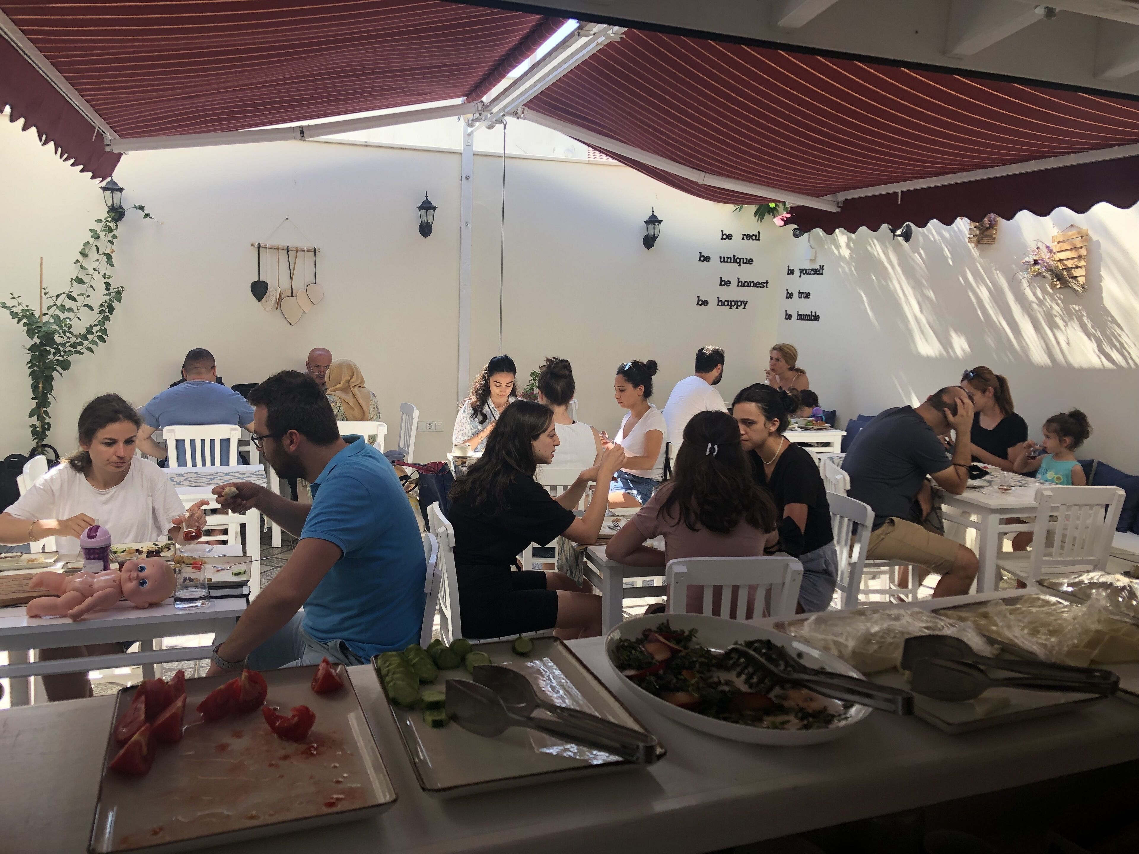 A group of people dining at an outdoor restaurant with white tables and chairs, surrounded by a covered area with a red roof.
