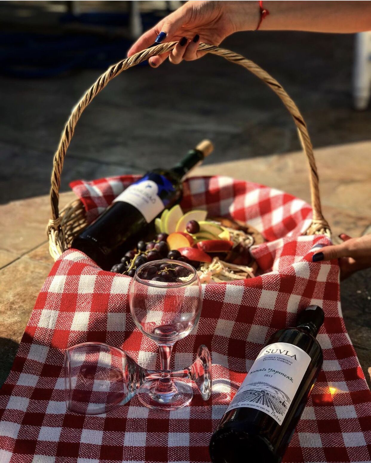 A picnic setup with a red and white checkered tablecloth, a wine bottle, a glass, and a basket containing food and a bottle of wine.