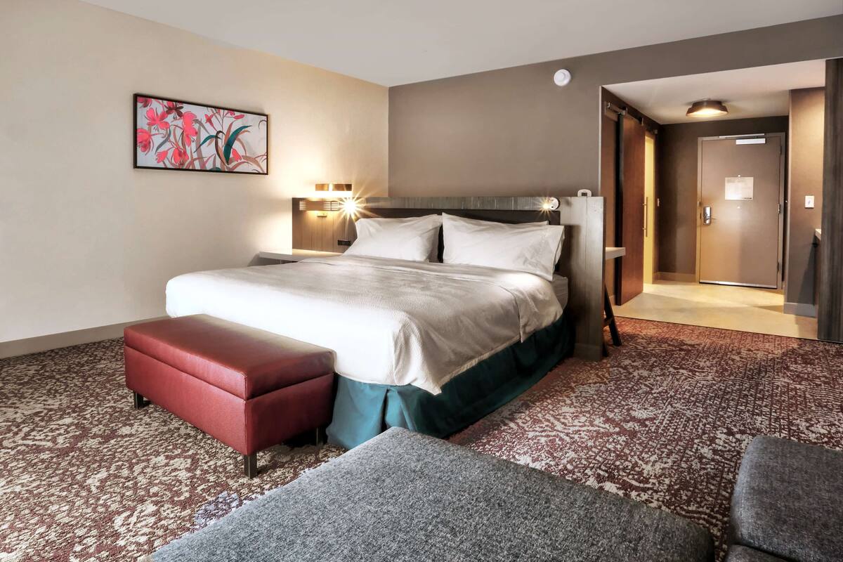 Hotellihuone, jossa on suuri sänky, punainen penkki, harmaa nojatuoli, kuvioitu matto ja taulu seinällä.