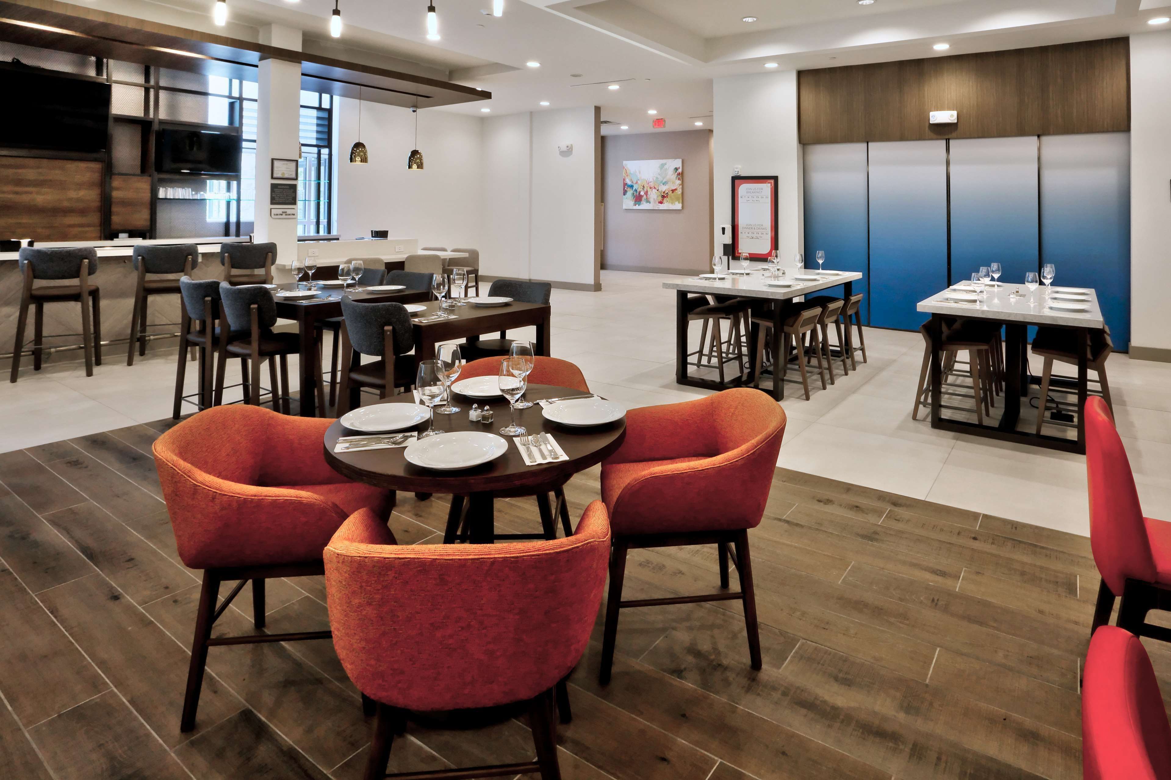 A modern dining area with round tables and red chairs, a bar area with stools, and a large blue accent wall.
